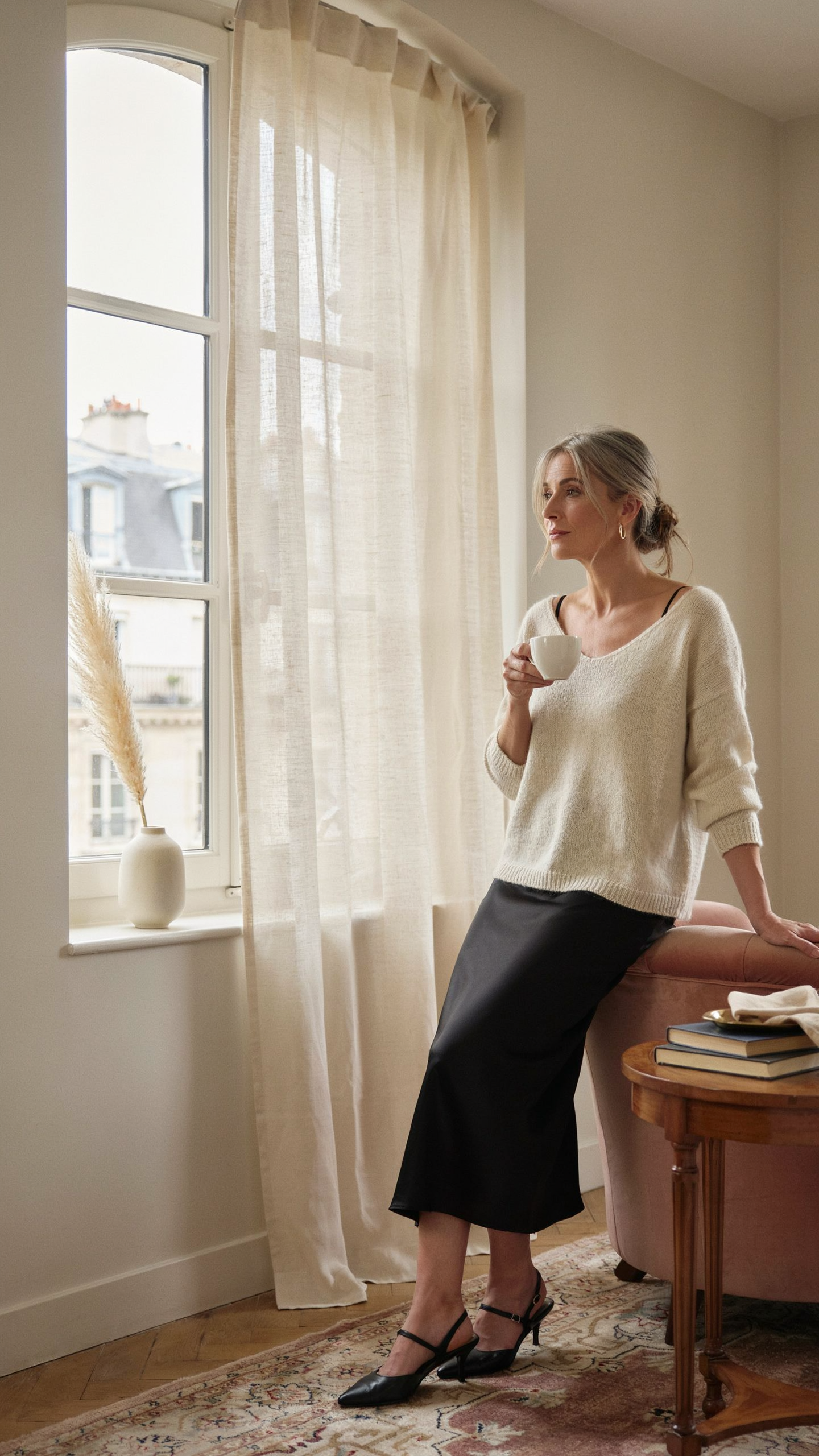 **Alt text:**
Woman in a cream knit sweater and black midi skirt sitting beside a small wooden table near a tall sunlit window with sheer curtains, holding a cup and looking thoughtfully into the distance in a calm, elegant room.
