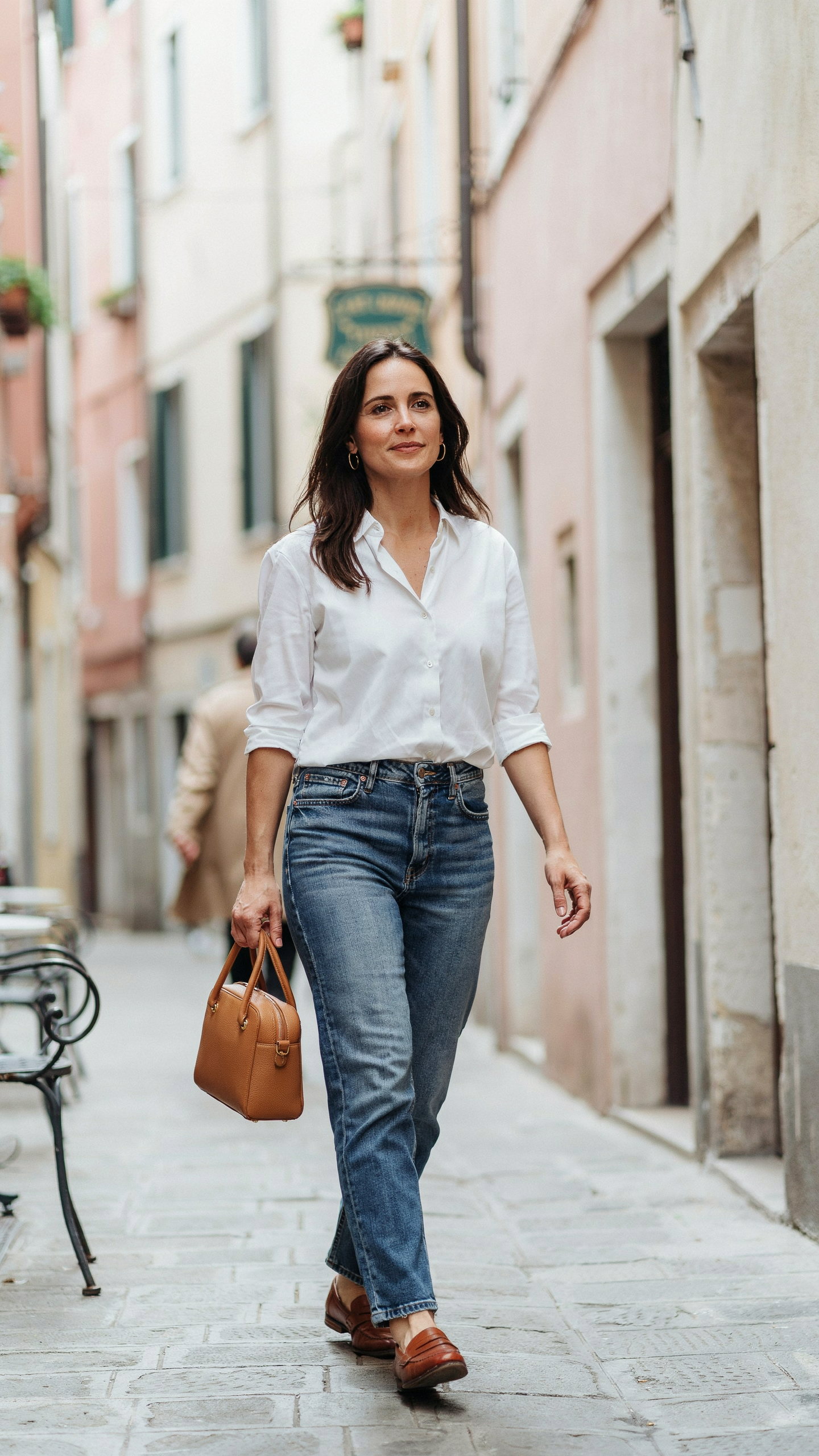 Woman wearing a crisp white button up shirt tucked into straight leg blue jeans with leather loafers, structured handbag, and gold hoop earrings.