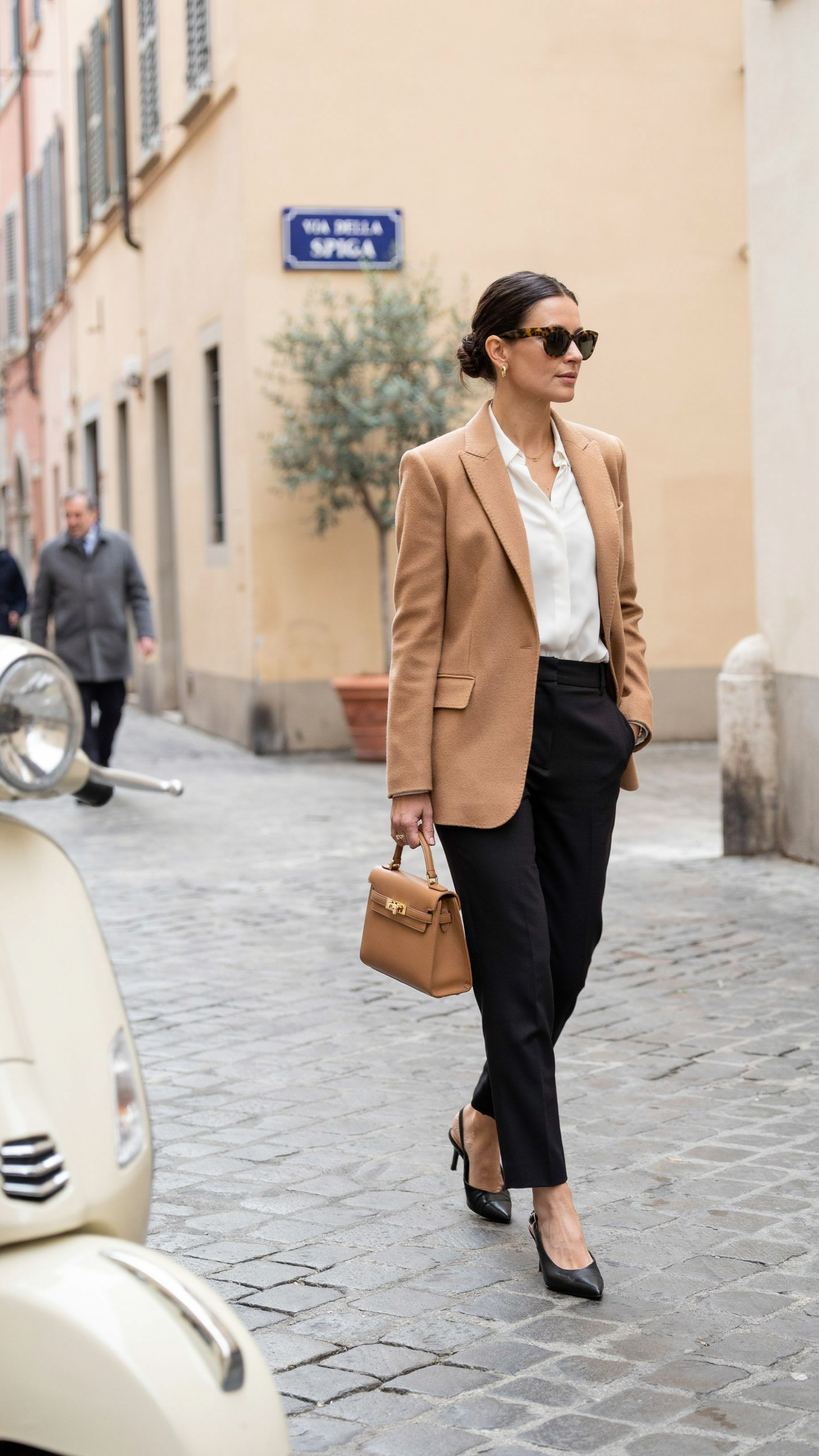 Elegant woman wearing oversized sunglasses with a tailored blazer, white shirt, leather handbag, and timeless Italian style clothing.